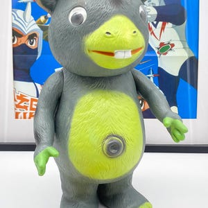 May include: A green and gray toy figure of a monster with a crown on its head. The toy has a large, toothy grin and a button on its belly. The toy is standing on a table in front of a poster with Japanese text.
