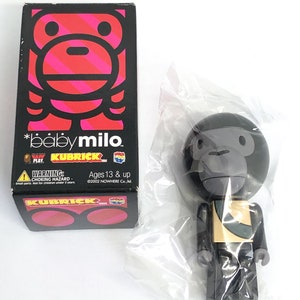 Peut inclure: Une figurine Baby Milo Kubrick noire et dorée dans un sac en plastique. La figurine porte une chemise dorée et noire et un pantalon noir. La figurine est debout sur une surface blanche. La boîte de la figurine est en arrière-plan. La boîte est noire avec un logo Baby Milo rouge et rose. La boîte porte le texte "*babymilo. PLAY KUBRICK Ages 13 & up ©2002 NOWHERE Co.,ltd."