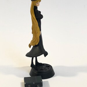May include: A black and gold anime figurine of a woman with long blonde hair wearing a black dress and hat. The figurine is standing on a black base with a circular design. A small black suitcase is in the foreground.