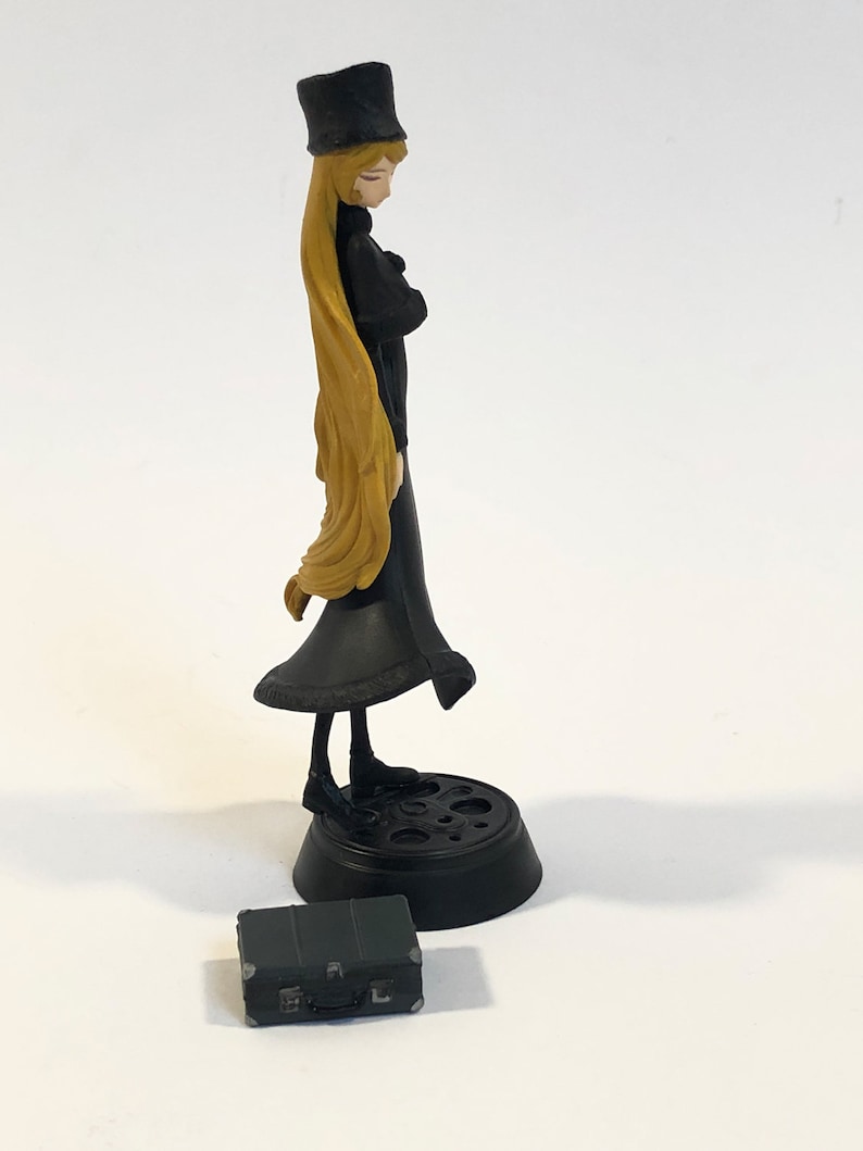 May include: A black and gold anime figurine of a woman with long blonde hair wearing a black dress and hat. The figurine is standing on a black base with a circular design. A small black suitcase is in the foreground.