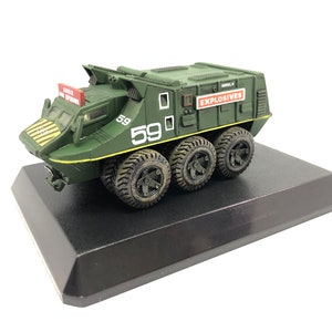 May include: A green toy military vehicle with the word "EXPLOSIVES" printed on the side. The vehicle has six wheels and the number "59" printed on the side. The vehicle is sitting on a black display base.