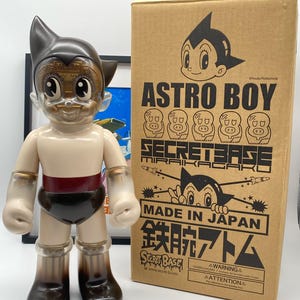 Könnte beinhalten: Eine transparente Plastikfigur von Astro Boy mit einem braunen Kopf und schwarzen Stiefeln. Die Figur steht auf einer braunen Oberfläche vor einer Box mit dem Text "Astro Boy Secret Base" und "Made in Japan".