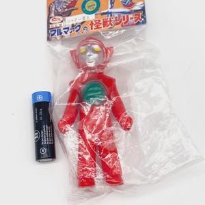 May include: A red Ultraman action figure in a plastic bag with a silver head and green accents. The packaging includes Japanese text and illustrations. A black and blue AA battery is next to the figure.