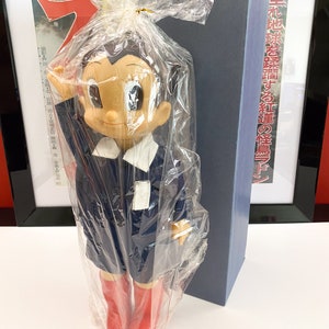 Peut inclure: Une poupée en plastique bleue et blanche d'Astro Boy, un personnage de manga japonais, portant des bottes rouges. La poupée est emballée dans du plastique transparent et se trouve à côté d'une boîte bleue avec du texte japonais.