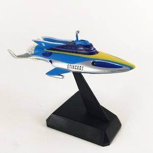 Peut inclure: Maquette du sous-marin Stingray, un engin aquatique futuriste avec des accents bleus, jaunes et argentés. Le modèle est monté sur un socle noir et porte l'inscription "STINGRAY" en lettres blanches.