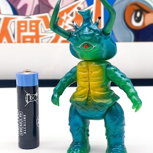 May include: A green and yellow plastic toy figure of a creature with a large head, four legs, and two long antennae. The figure is about 5 cm tall and is standing on a white surface.