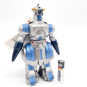 May include: A silver and blue robot toy with a drill on its head. The robot has a blue and silver body with a silver head and a drill on top. The robot is standing on a white surface.