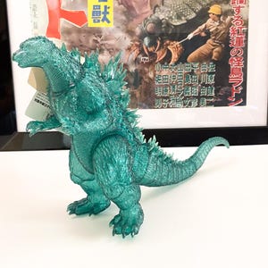 Peut inclure: Une figurine d'action Godzilla translucide verte avec une surface texturée. La figurine est posée debout, la bouche ouverte et la queue enroulée derrière elle. La figurine est placée devant une affiche avec du texte japonais.