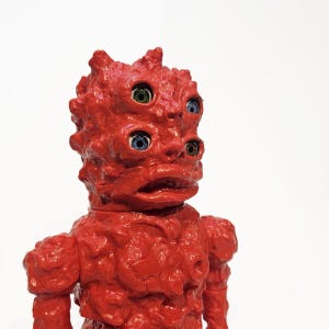 May include: A red, textured, humanoid figure with four eyes. The eyes are blue, green, and yellow. The figure has a large, open mouth.