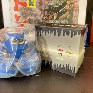 Puede incluir: Una figura de vinilo azul de un monstruo en una bolsa de plástico junto a una caja con un diseño en zigzag en blanco y negro. La caja tiene texto y logotipos japoneses. Un póster enmarcado está en el fondo.