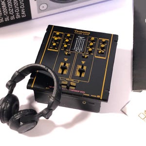 May include: A black and gold Technics DJ mixer with two channels, knobs, and faders. The mixer is on a white surface with a pair of black headphones.