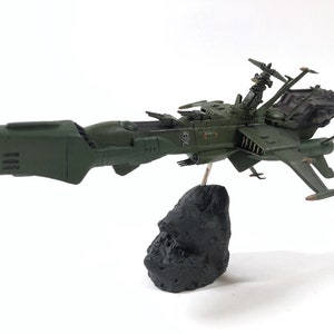 Puede incluir: Un modelo de nave espacial verde con una base negra. La nave espacial tiene un cuerpo largo y delgado con una nariz puntiaguda y un ala grande y plana a cada lado. La nave espacial está montada sobre una base de roca negra.