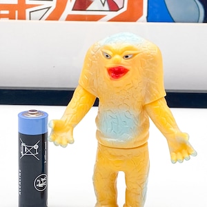 Peut inclure: Une figurine de monstre en plastique jaune et bleu avec une grande bouche et une surface texturée. La figurine est debout sur une surface blanche.