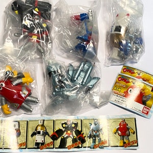 Puede incluir: Un conjunto de seis juguetes robots en bolsas de plástico individuales. Los robots son plateados, rojos, azules y negros. Los juguetes forman parte de una colección de juguetes robots. El embalaje incluye una tarjeta con el texto "25th Anniversary" y "Super Robot Taisen OG", y una imagen de un robot rojo.