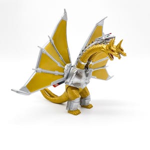 Peut inclure: Une figurine d'action King Ghidorah en or et argent avec trois têtes et de grandes ailes jaunes. La figurine est posée avec ses ailes déployées et ses têtes regardant vers l'avant.