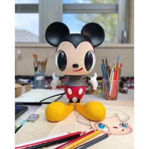 Peut inclure: Figurine de Mickey Mouse avec de grands yeux expressifs, des oreilles noires et une tenue rouge et blanche. La figurine a des chaussures jaunes et des gants blancs. L'arrière-plan comprend des fournitures d'art et un dessin.