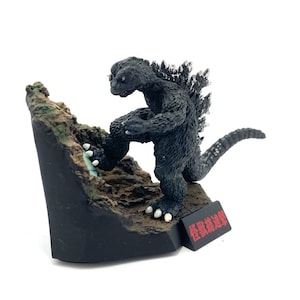 Peut inclure: Figurine noire de Godzilla grimpant un terrain rocheux. La figurine a une surface texturée, des griffes blanches et une nageoire dorsale épineuse. La base est noire avec du texte japonais.