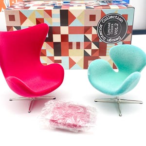 Op de afbeelding: Twee miniatuur eivormige stoelen, een roze en een lichtblauw, met een roze kussen op de voorgrond. De stoelen staan op een wit oppervlak en een doos met de tekst "Design Interior Collection Vol.2 Limited Colors" is op de achtergrond.