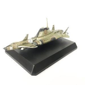 Könnte beinhalten: Ein Modell eines futuristischen Flugzeugs, hauptsächlich olivgrün mit roten und schwarzen Akzenten. Das Flugzeug ist auf einem schwarzen rechteckigen Sockel montiert. Das Design umfasst Flügel, einen zentralen Körper und eine Finne.