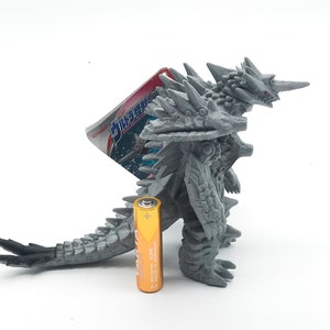 May include: A gray plastic toy monster with spikes and red eyes. The monster is standing on a white surface with a battery next to it. The battery is gold and has the text "Energizer" and "MAX" on it.