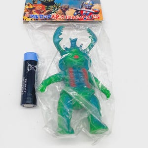 May include: A green and blue toy monster figure in a plastic bag, with red accents on its chest. A blue alkaline battery is next to the toy. A colourful card with Japanese text is at the top.
