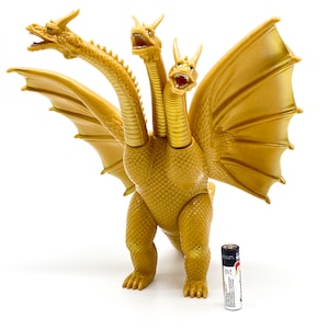 Peut inclure: Un jouet de dragon doré à trois têtes avec des ailes. Le jouet est posé sur une surface blanche avec une pile pour l'échelle.