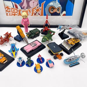 Puede incluir: Una colección de vehículos y figuras de juguete vintage, que incluye coches, aviones y un helicóptero. Los juguetes son de varios colores, como rojo, amarillo, azul y verde. Una imagen enmarcada está en el fondo, y una muñeca está de pie cerca de los juguetes.