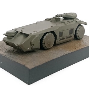 May include: A green military-style toy vehicle with a turret on top, sitting on a brown base.