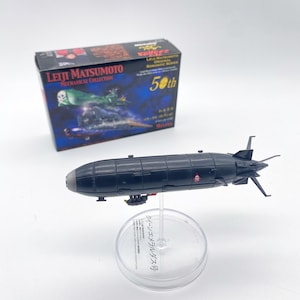 Peut inclure: Un modèle de vaisseau spatial noir et rouge avec un support en plastique transparent. Le support porte le texte "クイーンエメラルダス号" en japonais. La boîte derrière le vaisseau spatial est bleue et porte le texte "LEIJI MATSUMOTO MECHANICAL COLLECTION 50th 9nres".