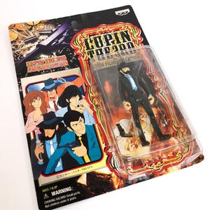Peut inclure: Figurine LUPIN THE 3RD dans son emballage d'origine. La figurine représente un homme en costume noir, chapeau et barbe. L'emballage présente des illustrations de dessins animés et le texte "LUPIN THE 3RD". L'emballage est noir, rouge et or.