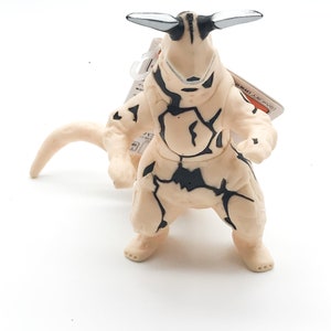 May include: A beige and black monster toy with a cracked texture and two white antennas. The toy has a long tail and is standing on a white surface.