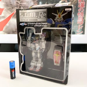 以下が含まれることがあります： 「S.C.M.Re SPECIAL CREATIVE MODEL IMPULSE GUNDAM」と「スペシャルクリエイティブモデル リアルエディション」と書かれた銀色のラベルが付いた黒い箱。箱の中には、白と青の小さなプラスチック製のロボットモデルが入っています。