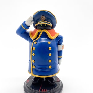 Peut inclure: Figurine bleue représentant un personnage en uniforme militaire saluant. La figurine a un chapeau bleu, un manteau avec des boutons dorés et un col orange. La base est noire avec des accents rouges. Le personnage a des yeux verts.
