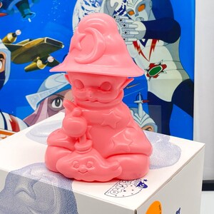 Peut inclure: Figurines en plastique rose d'un personnage portant un chapeau avec une lune croissante et une étoile. Le personnage tient un bâton et a une étoile sur la poitrine. La figurine est assise sur une boîte blanche avec un motif bleu et blanc.