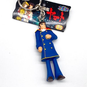 Peut inclure: Un porte-clés bleu avec une figurine d'un homme en manteau bleu à boutons dorés et pantalon bleu. Le porte-clés a un anneau argenté et une étiquette avec du texte et des illustrations japonaises.
