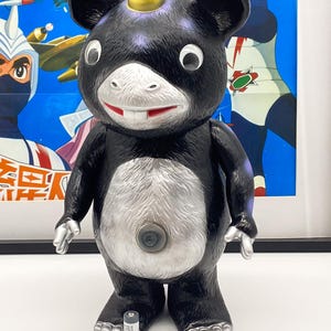 May include: Black and silver toy figure with a gold crown on its head. The figure has a large, round belly and small, stubby legs. It is standing on a white surface.