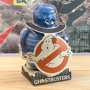 Op de afbeelding: Een blauw, glinsterend Stay Puft Marshmallow Man-figuur met een doorzichtige hoed en kraag. Het figuur heeft een Ghostbusters-logo aan de voorkant en staat op een doos met de tekst "GHOSTBUSTERS".