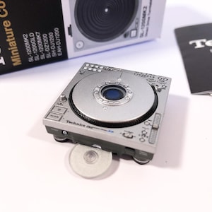Puede incluir: Una réplica de platina Technics SL-1200MK2 plateada, un modelo en miniatura de un equipo de DJ clásico. La platina tiene un plato plateado con un centro negro y un panel de control negro con letras blancas. La platina está sobre una superficie blanca.