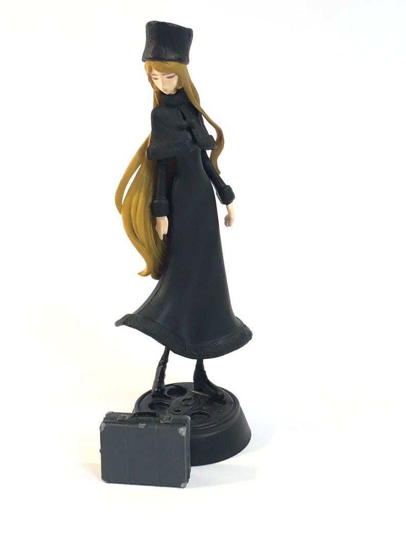May include: A black and gold figurine of a woman wearing a black coat and a black hat. She is holding a black briefcase.