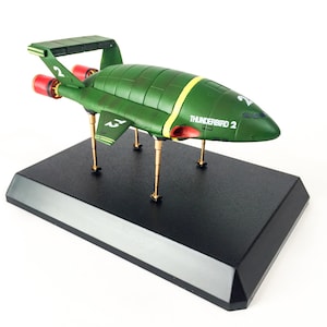 Puede incluir: Un modelo verde del avión Thunderbird 2, con rayas amarillas y detalles de motor rojos, se exhibe sobre una base negra. El modelo tiene el número "2" en las alas y el texto "THUNDERBIRD 2" en el lateral. El modelo está sostenido por patas doradas.