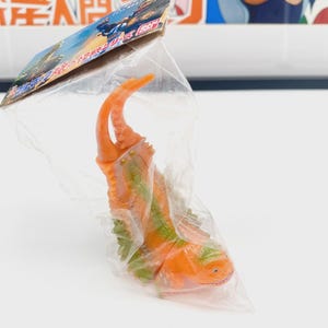 May include: An orange and green toy creature in a clear plastic bag. The toy has a claw-like feature and a small head. The packaging has Japanese text and a colourful illustration.