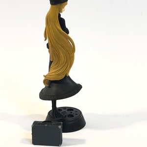 May include: A black and gold figurine of a woman wearing a black dress and a black hat. She is holding a black briefcase in her hand. The figurine is standing on a black base.
