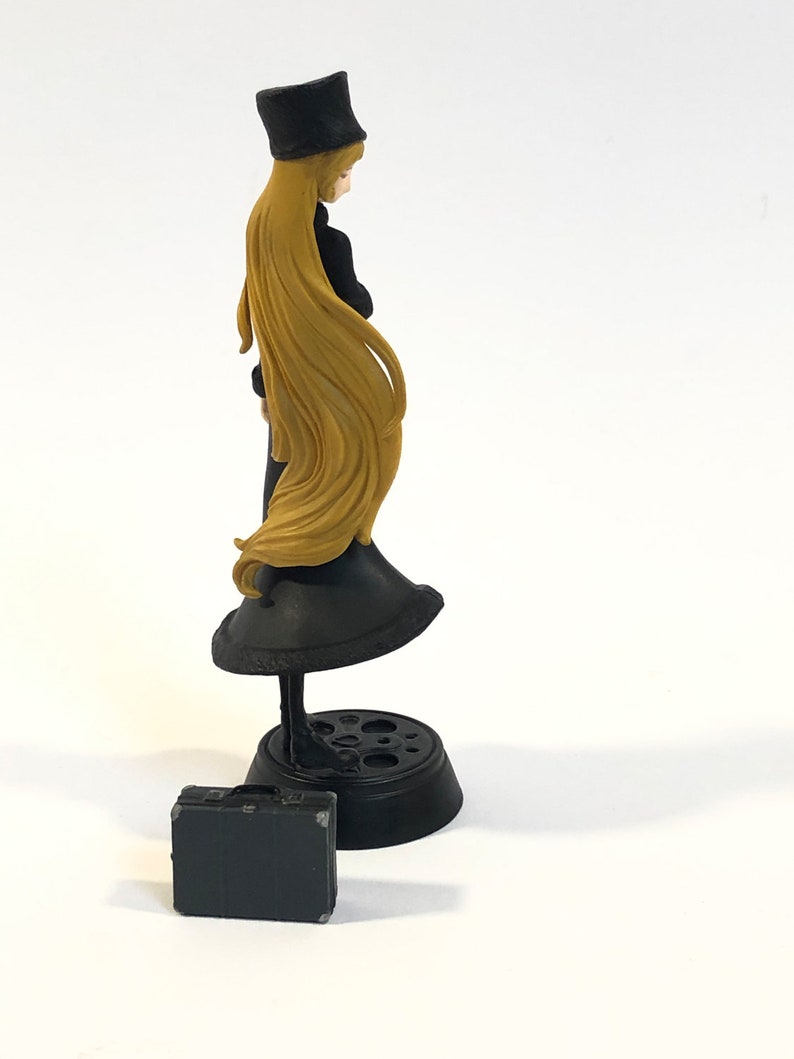 May include: A black and gold figurine of a woman wearing a black dress and a black hat. She is holding a black briefcase in her hand. The figurine is standing on a black base.