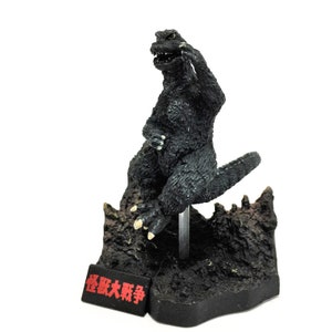 Peut inclure: Figurine noire de Godzilla, posée sur un socle rocheux. La figurine a une texture détaillée et est montée sur un support transparent. Un petit panneau rouge avec du texte japonais est à la base. La figurine mesure environ 13 cm de haut.
