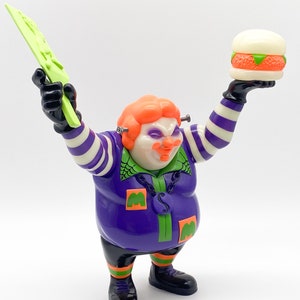 May include: A purple and green toy figure of a clown holding a green knife and a white burger. The clown has orange hair, a green shirt, and a purple vest with a chain and a silver pendant. The clown is wearing black pants and black shoes.