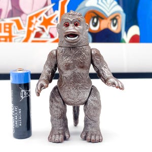 Pode incluir: Uma figura de brinquedo de gorila marrom e prateado com a boca aberta e olhos vermelhos. A figura está em pé sobre uma superfície branca.