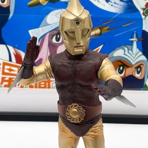 Peut inclure: Une figurine d'action dorée et brune d'un super-héros portant un casque avec un joyau vert. La figurine a une ceinture dorée avec un motif circulaire et tient une arme dans chaque main.
