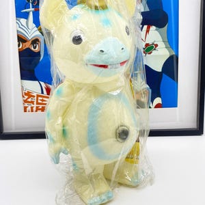 May include: A white and blue plush toy with a gold crown. The toy has a blue nose and mouth, and a single black eye. The toy is wrapped in plastic.