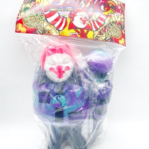 Peut inclure: Un clown en vinyle violet, bleu et noir avec une perruque rose et un visage blanc. Le clown tient un microphone violet. Le jouet est emballé dans un sac en plastique transparent avec une étiquette colorée qui dit "Evil Mo" et "Ron English".