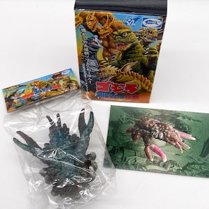 May include: A green and brown plastic toy monster from the Godzilla franchise. The monster is in a plastic bag with a card showing a picture of the monster. The card has the text "Godzilla" and "Ultraman" on it.